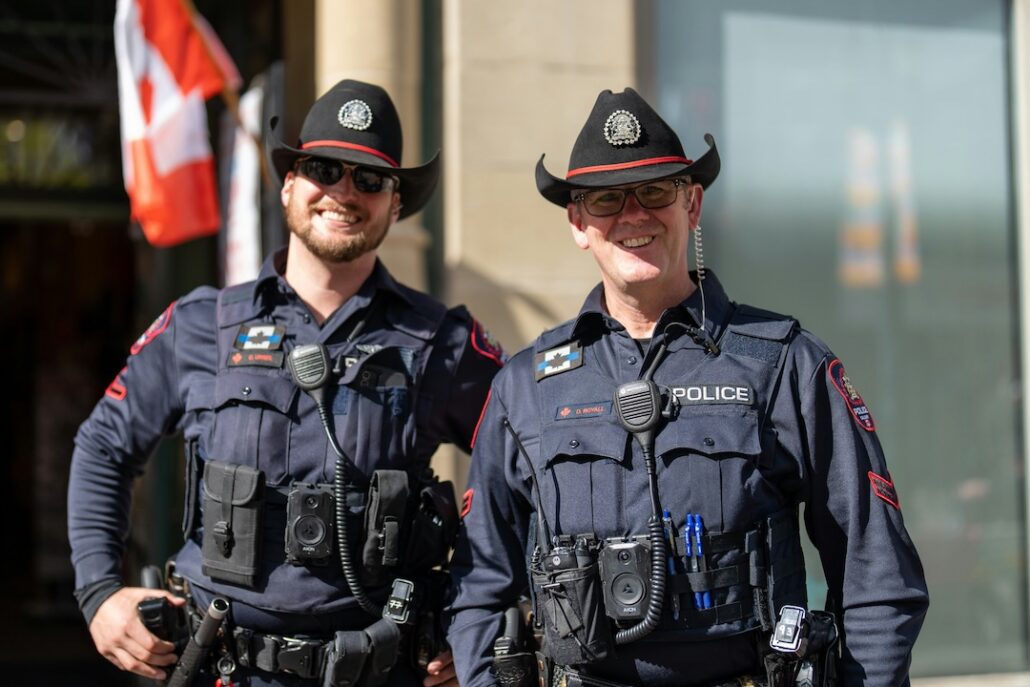 Two police officers in dark uniforms and cowboy hats smile at the camera outdoors with gear and radios visible on their vests